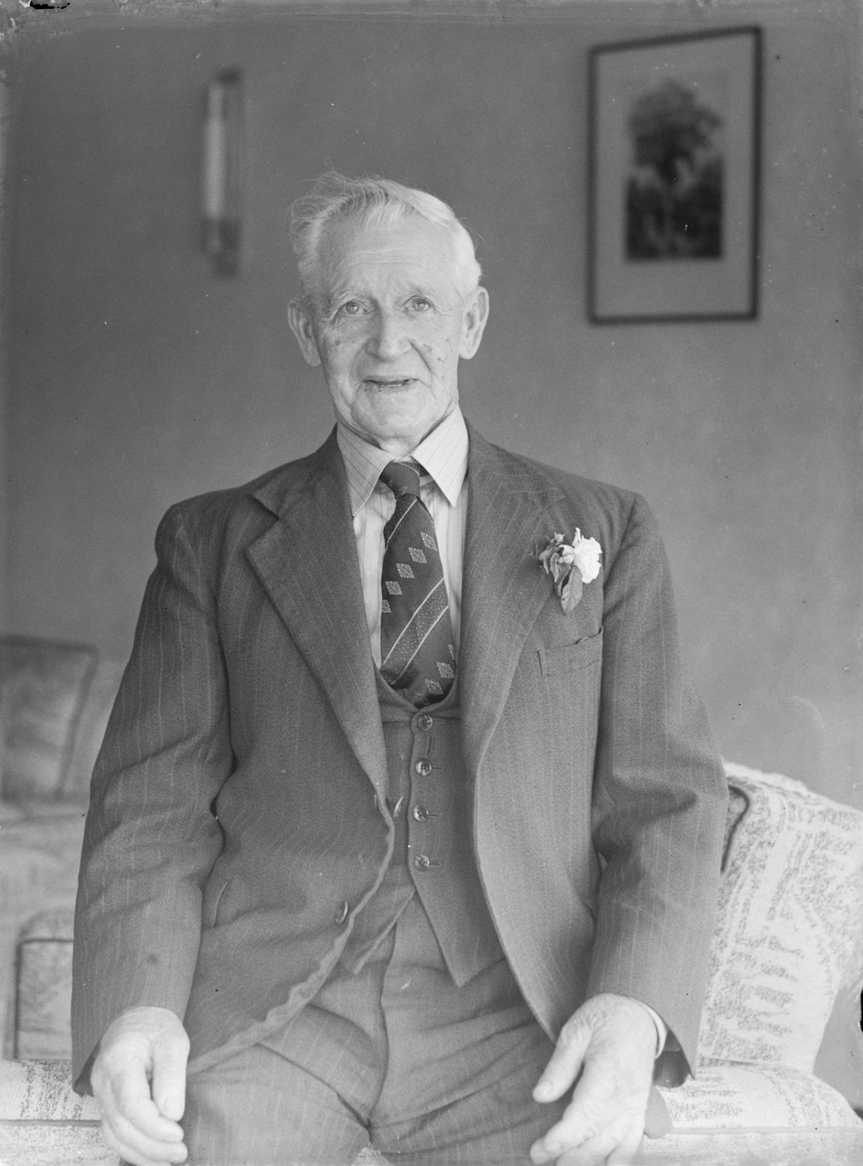 Elderly man seated in an interior setting, wearing a pinstriped suit in an armchair